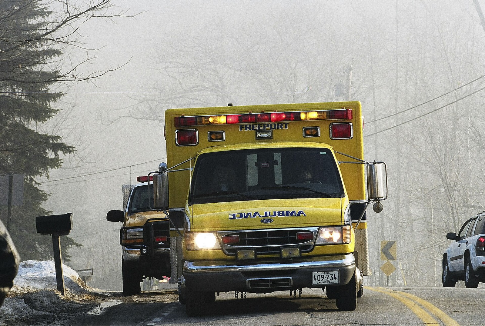 A yellow Freeport, Maine municipal ambulance with its lights on, parked on a foggy two-lane rural road, a fire department pickup behind it.