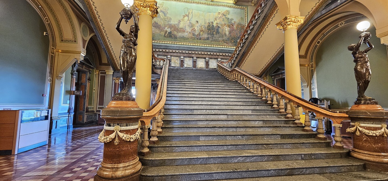 A wide marble staircase with bronze statues on the newel posts and a mural above, inside the Iowa State Capitol.